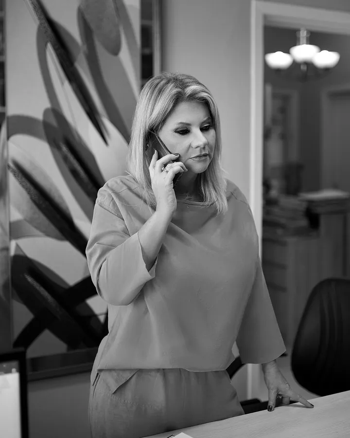 Luciana Meirelles ao telefone, em pé junto à mesa do escritório